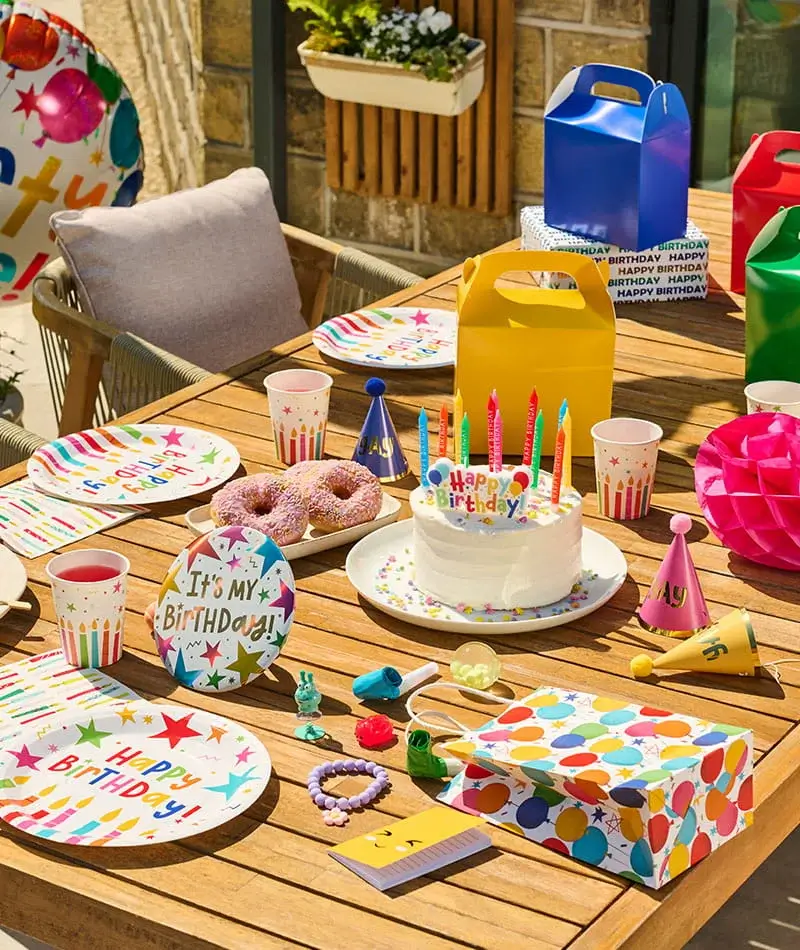 Colourful birthday party table with a birthday cake, party hats, gift boxes, drinks, doughnuts, and festive tableware for a children's celebration. Colourful birthday party table with a birthday cake, party hats, gift boxes, drinks, doughnuts, and festive tableware for a children's celebration.